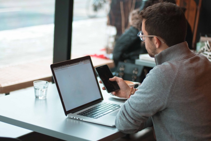 A man using his laptop and smartphone while sitting at a table in a cafe.