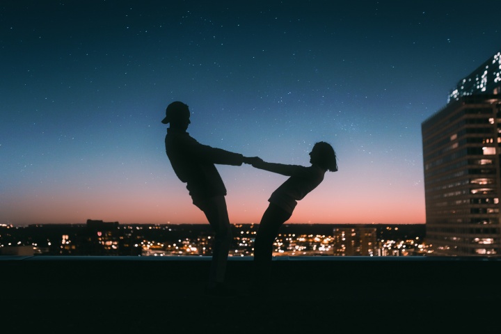a man and woman in silhouette against a city skyline and starry sky, holding hands and leaning back mid-twirl