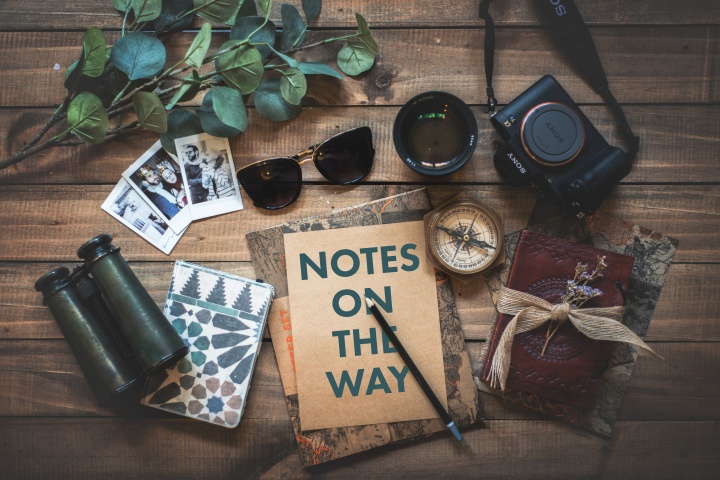 An assortment of items flat laid on a table, including a camera, a notebook, and sunglasses