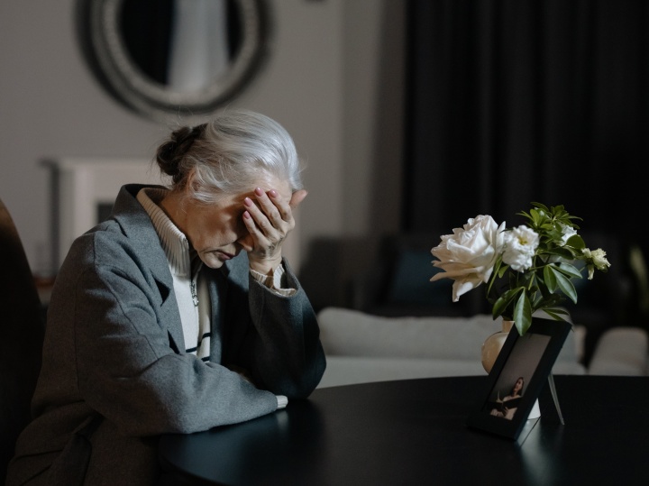 a woman with her head in her hand as she sits at a table across from white flowers and a photo frame