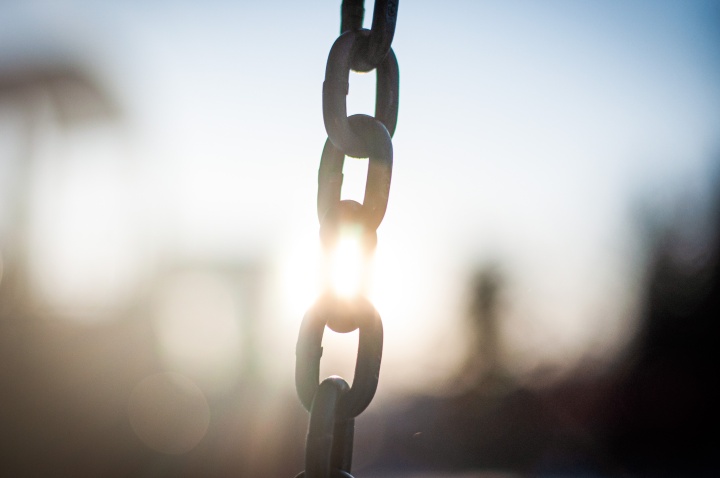 a length of chain in the foreground with the sunset shining in the background, the light hitting the chain in the center, interrupting the chain's solid appearance