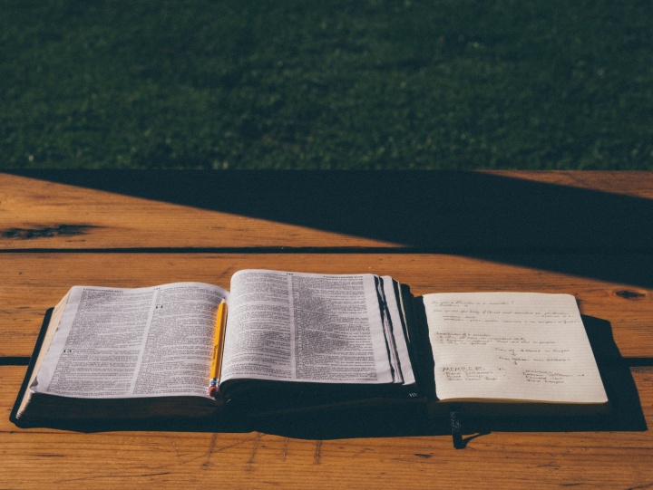 A opened Bible laying on a picnic table beside a notebook.