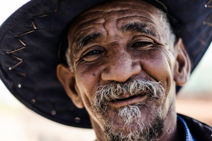 Upclose photo of a older man with smile and grey goatee - wearing a hat.