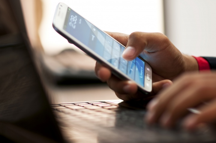 A person typing on a laptop and check their smartphone's homescreen.