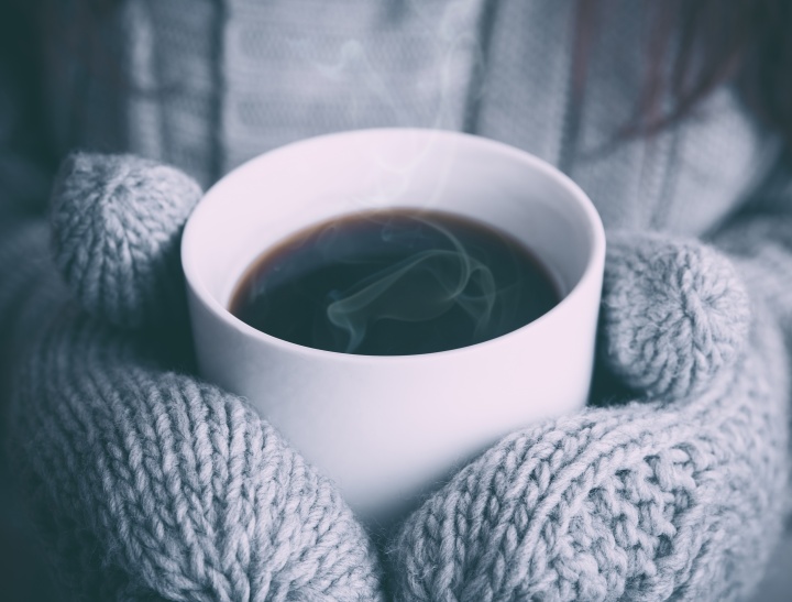 A person wearing mittens holding a hot beverage with steam rising.