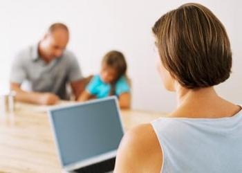 Woman working on laptop with husband and child in background - The Juggling Act