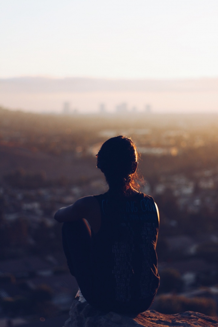 A woman sitting by herself on a cliff looking out over the town. 