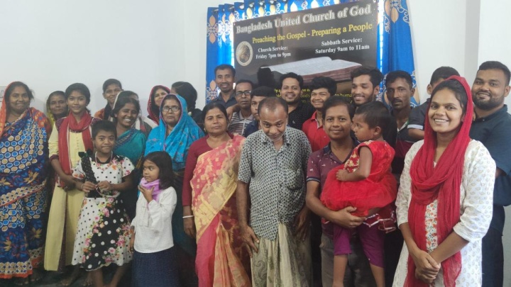 a group of brethren standing indoors with a Bangladesh UCG banner in the background