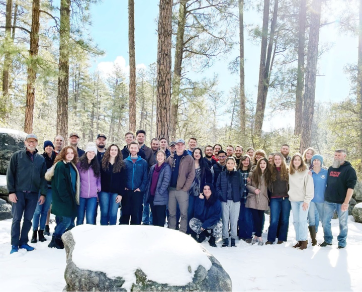 A group of young adults gathered outdoors in the snow with trees in the background