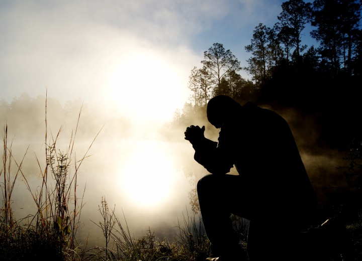 A man praying outside by a field lit by the morning sun.