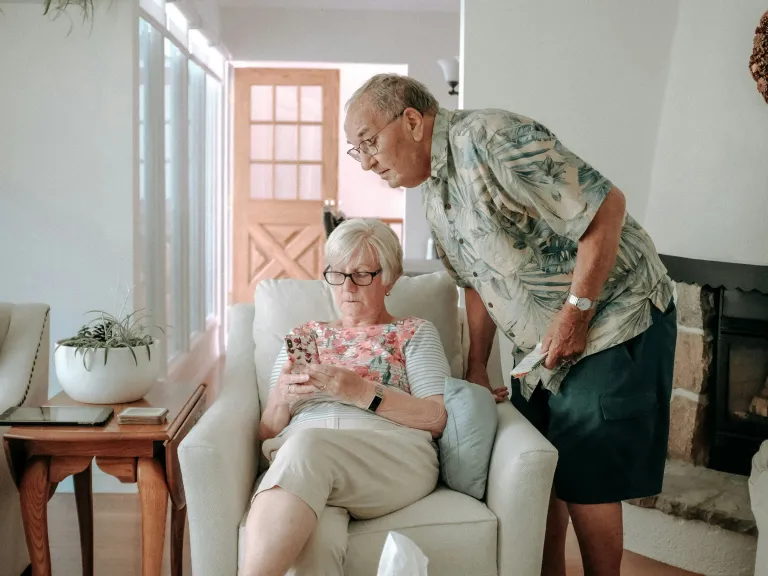 a woman seated in an armchair with a man leaning over to look at what she's showing him on her phone