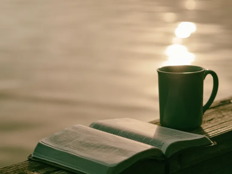 The Sabbath is a time for a Christian to study God’s word and spend extra time in prayer and meditation. An open Bible and a ceramic mug sitting on a table with a pond in the background.