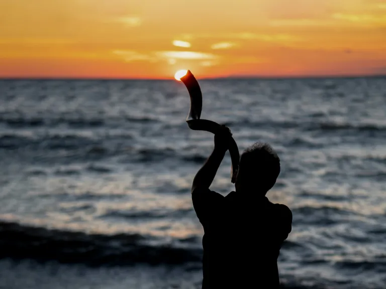 Feast of Trumpets a silhouette of a person blowing into a shofar with sunset over the ocean in the background