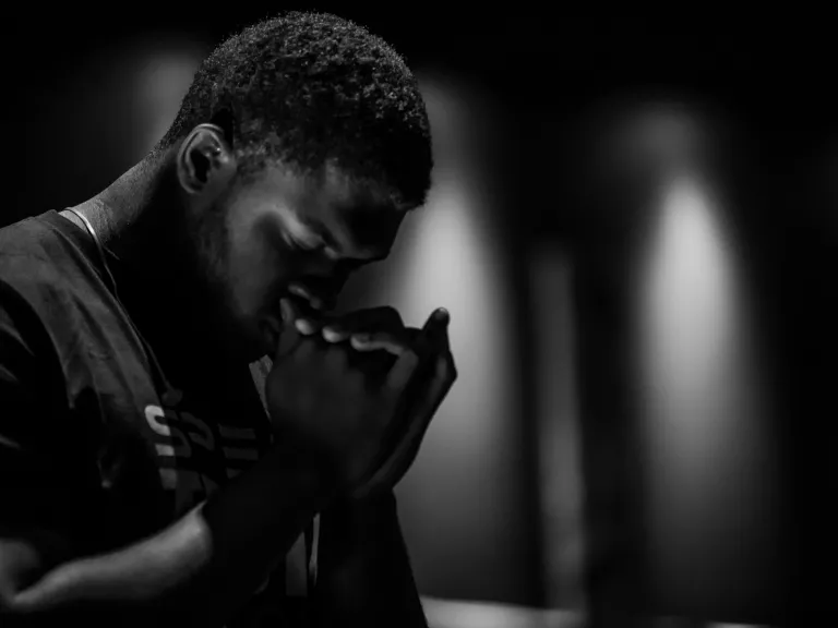 As dedicated Christians, we are committed to praying daily. a man kneeling in prayer with folded hands in a dark room