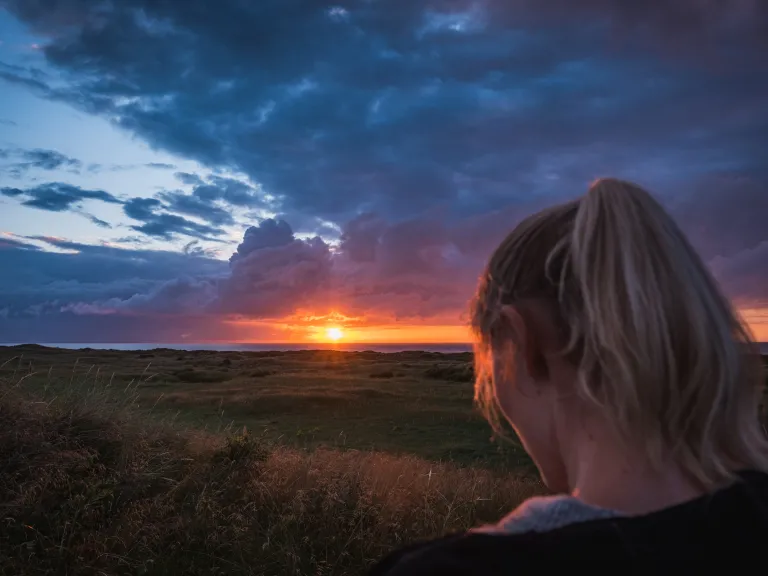 Never underestimate the power of God to finish His perfect work in you. Christ is preparing places for His followers in His Kingdom. A woman in a field with the sun setting in the distance.