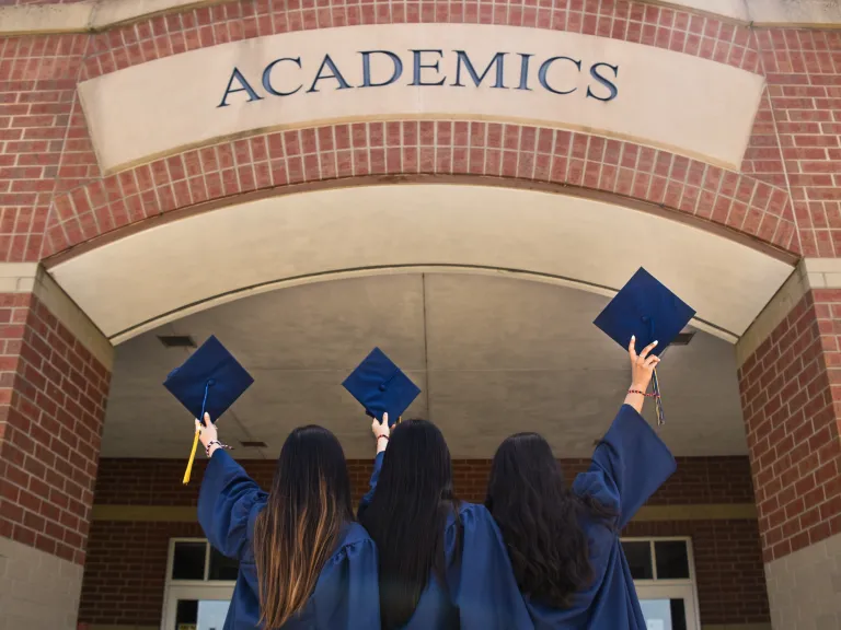 It's time to send in your graduation announcement! three women in blue graduation robes and holding graduation caps under a brick archway
