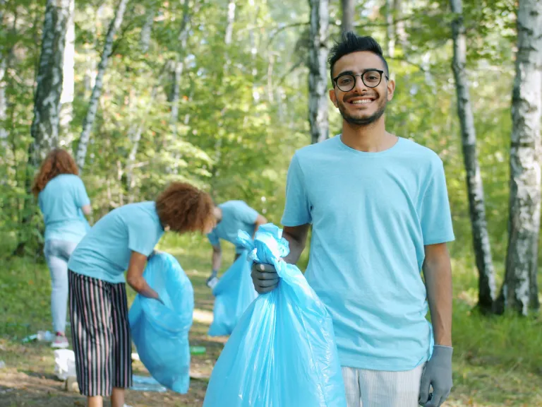 a man holding up a plastic trash bag while a team behind him cleans up trash in the forest