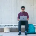 a man sitting ona bench and looking at a laptop with a backpack on the ground beside him