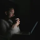 a woman looking at the laptop screen in a dark room and holding a coffee mug close to her face