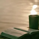An open Bible and a ceramic mug sitting on a table with a pond in the background.