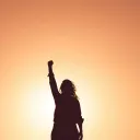 a woman's silhouette with a triumphant raised fist against a golden orange sky