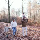 A mom and dad walking in the woods with their two kids. The daughter is on the shoulders of the dad and the son is holding hands with the mom.