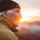 an elderly man wearing a hat and coat as he stands outdoors looking into the distance with a sunset in the background