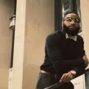 a well-dressed man standing in front of a concrete building and leaning on a metal stair rail