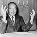 Rev. Martin Luther King, head-and-shoulders portrait, seated, facing front, hands extended upward, during a press conference.