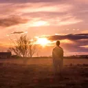 A man standing in a pasture looking at the setting sun.