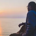 A man sitting by a body of water looking at the sunset.