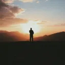 A silhouette of a person stand on a hill with the sun in the background.