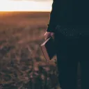 A person standing in a field holding a Bible while the sun is setting.