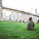 People sitting at a park by a large bridge.