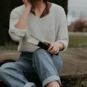 A girl in cropped mom jeans sits on the ground with a Bible in her lap. A wide brim hat sits next to her and she has a flowery collared shirt on under a white sweater.