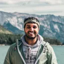 a man stands outdoors with mountains and a lake behind him