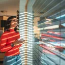 a woman standing indoors and looking out through a reflective window while holding a coffee cup