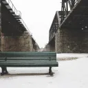 A park bench surrounded by snow and underneath a bridge.