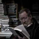 An older man with a beard reading a book. Stacks of books are behind him.
