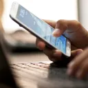 A person typing on a laptop and check their smartphone's homescreen.