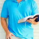 Man reading a Bible while leaning against a wall.