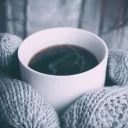 A person wearing mittens holding a hot beverage with steam rising.