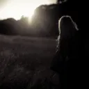 A woman standing in a field with the sun glaring behind a row of trees.