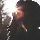 A girl wearing a jacket and hood looking at the camera with sunrays behind in head.