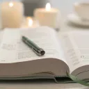 a bible with a pen lying across its open pages and candles and a teacup in the background