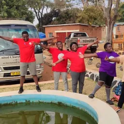 a group of children standing outside by a pool with the Church bus parked behind them