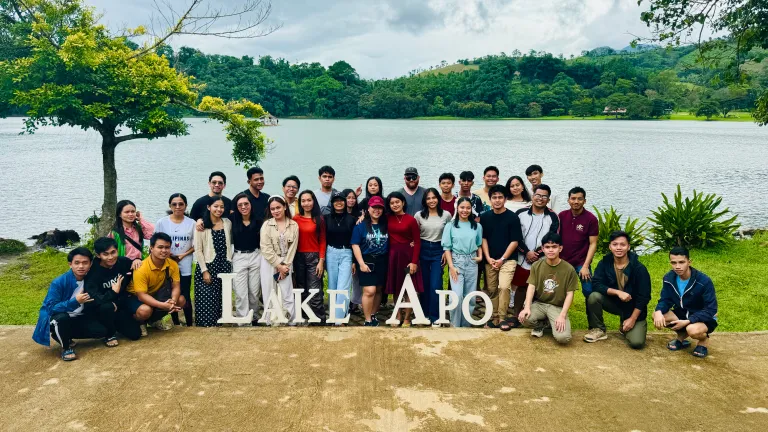 a group of people standing in front of a lake with a sign &quot;Lake Apo&quot; in front of them