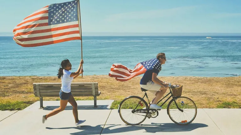 Man on bicycle and girl running behind with American flags.