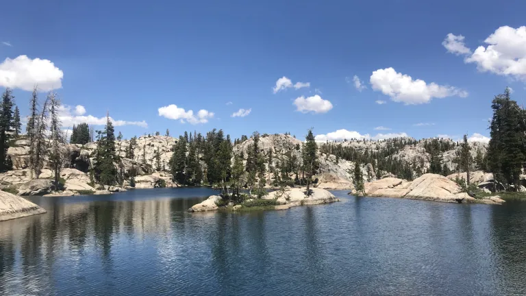 a lake with rocks and trees surrounding its perimeter and a blue sky with clouds above