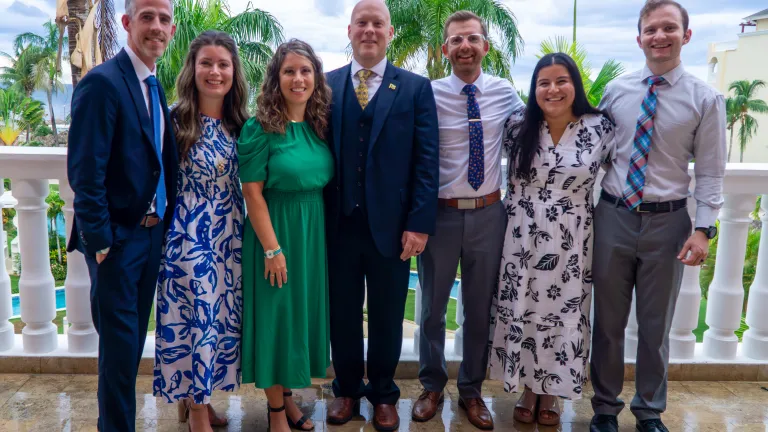 seven people standing outside on a balcony with palm trees behind them